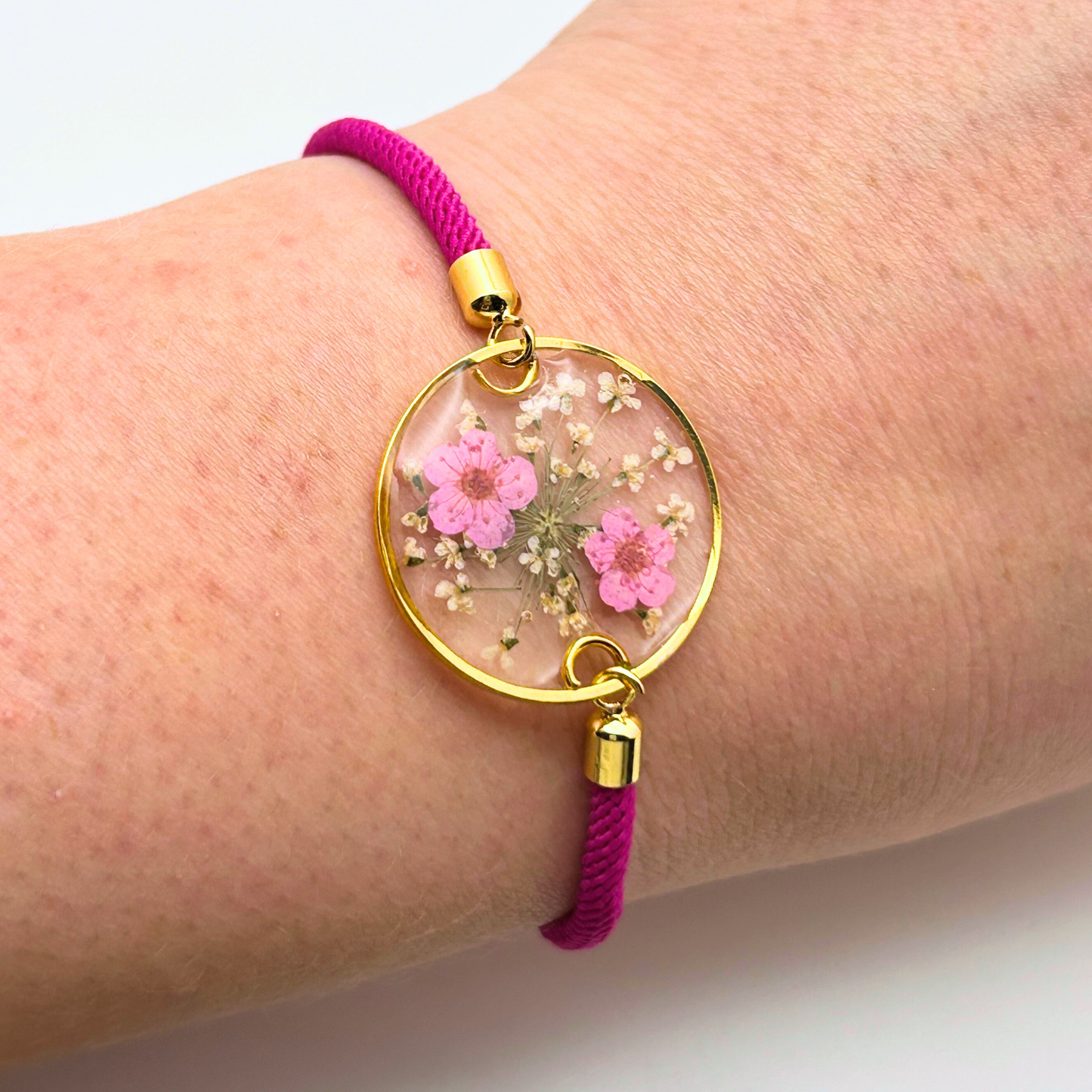 A gold-toned bracelet with a clear, circular pendant containing pink flowers and white accents. The pendant is attached to a fuchsia-colored cord. The bracelet is a delicate piece of jewelry.