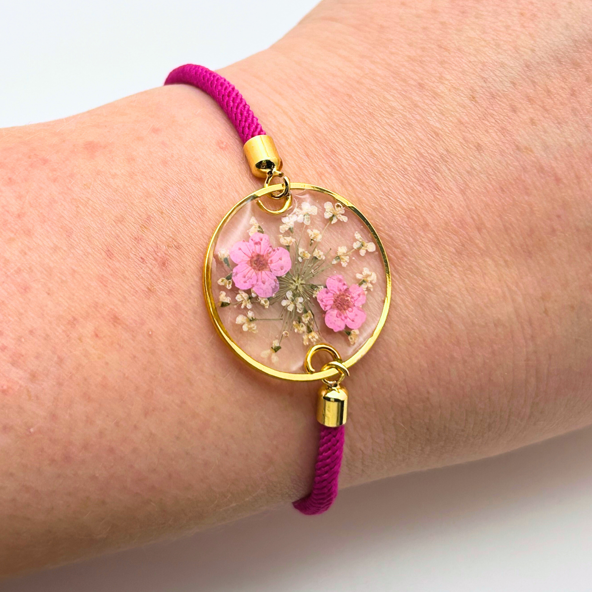 A gold-toned bracelet with a clear, circular pendant containing pink flowers and white accents. The pendant is attached to a fuchsia-colored cord. The bracelet is a delicate piece of jewelry.