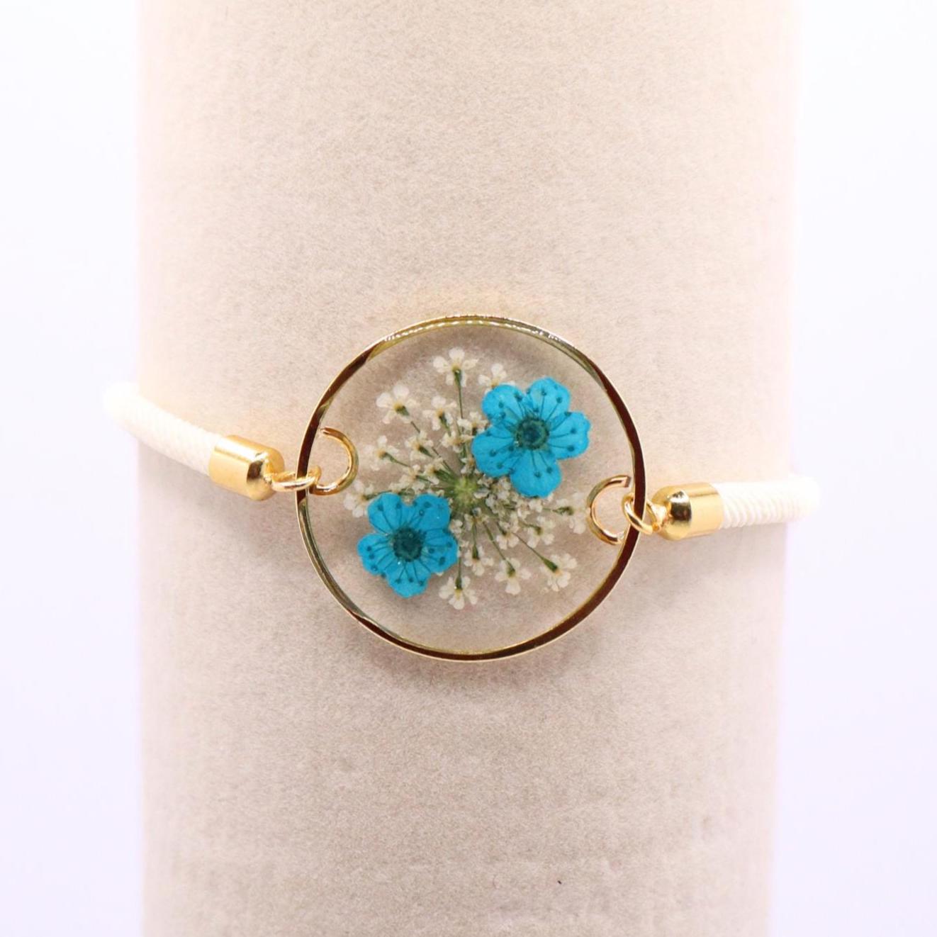 Pressed blue flower resin bracelet with gold bezel on white adjustable cord displayed on jewelry stand