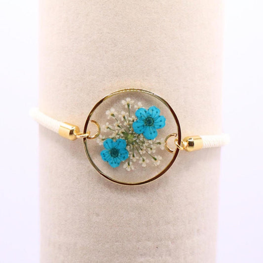Pressed blue flower resin bracelet with gold bezel on white adjustable cord displayed on jewelry stand