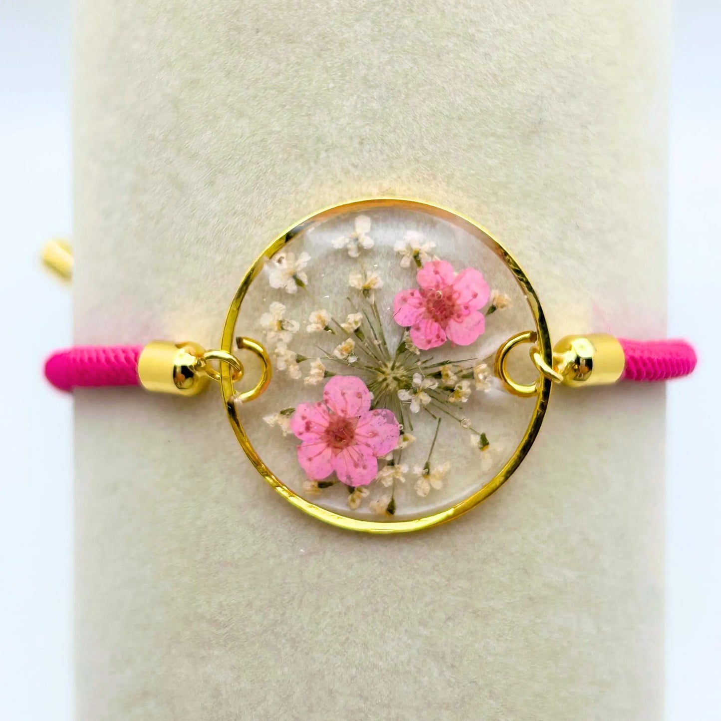 A gold-toned bracelet with a clear, circular pendant containing pink and white dried flowers in resin. The bracelet has a bright pink cord and gold-colored metal accents.