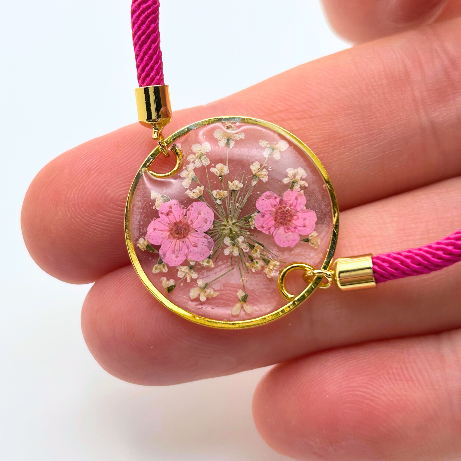 A round pendant bracelet with a gold-colored frame, filled with pink and white dried flowers in resin. The pendant is attached to a bright pink cord with gold-colored metal accents.