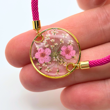 A round pendant bracelet with a gold-colored frame, filled with pink and white dried flowers in resin. The pendant is attached to a bright pink cord with gold-colored metal accents.