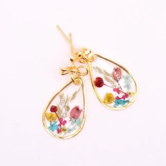 A pair of gold-colored earrings with teardrop-shaped pendants. Each pendant features a clear resin encasing dried flowers in shades of red, pink, yellow, and blue. The earrings are finished with an 18k gold-plated bow posts