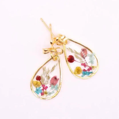 A pair of gold-colored earrings with teardrop-shaped pendants. Each pendant features a clear resin encasing dried flowers in shades of red, pink, yellow, and blue. The earrings are finished with an 18k gold-plated bow posts