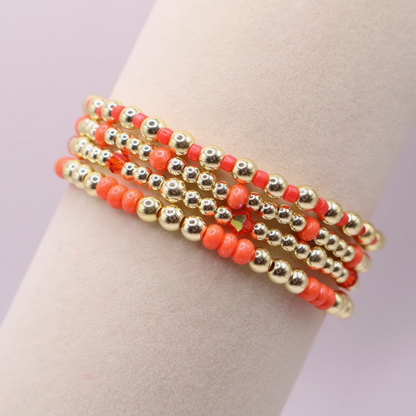 Stack of handmade stretch bracelets featuring vibrant coral Preciosa seed beads, faceted crystal accents, and polished gold-tone hematite beads on a clean white background.