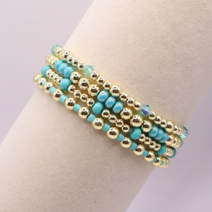 Layered bracelet stack with turquoise Preciosa seed beads, shimmering crystal accents, and gold-tone hematite beads on a clean white background.