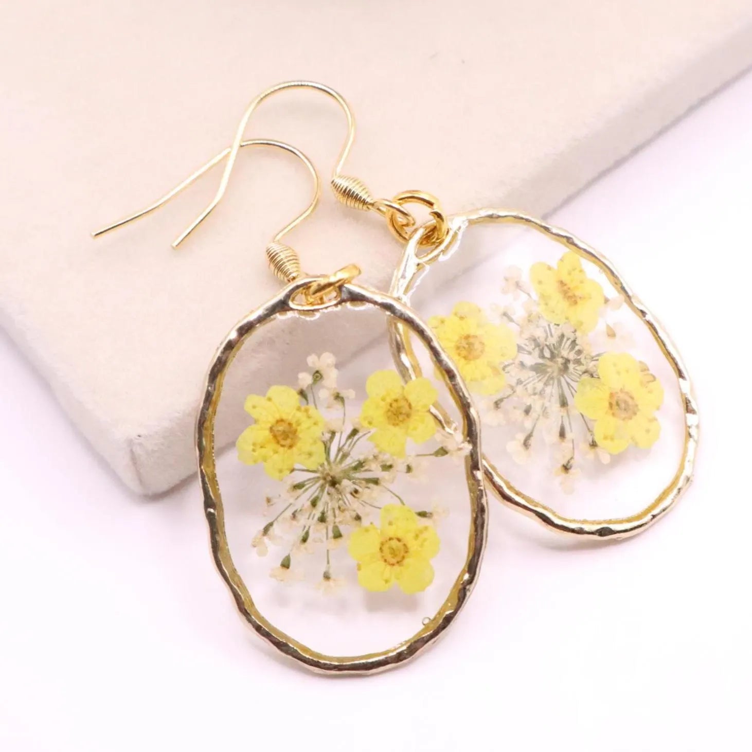 Close up of handmade resin earrings with pressed yellow flowers and fine white floral accents in clear oval shape with gold trim and hook ear wires